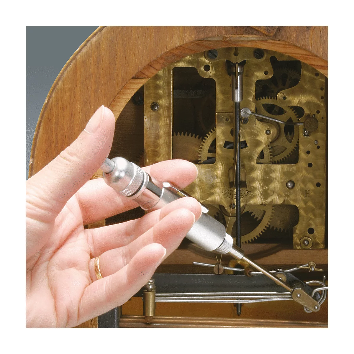 Person hält Werkzeug an Uhrwerk. Goldene Zahnräder und Holzrahmen.