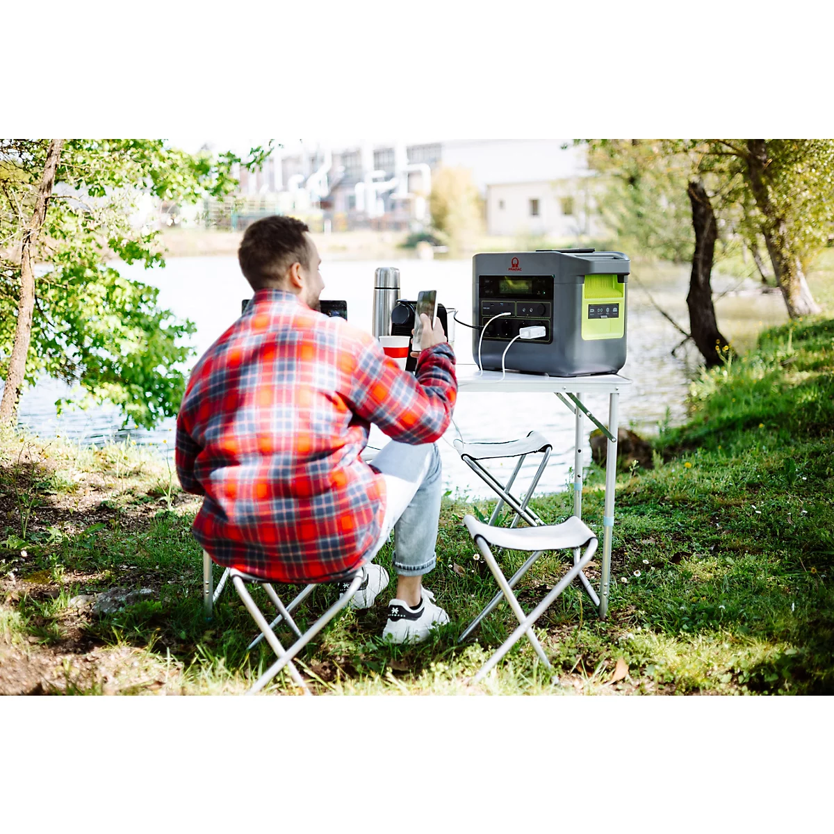 Ein Mann sitzt mit einem Smartphone an einem Campingtisch am See und nutzt eine tragbare Powerstation.