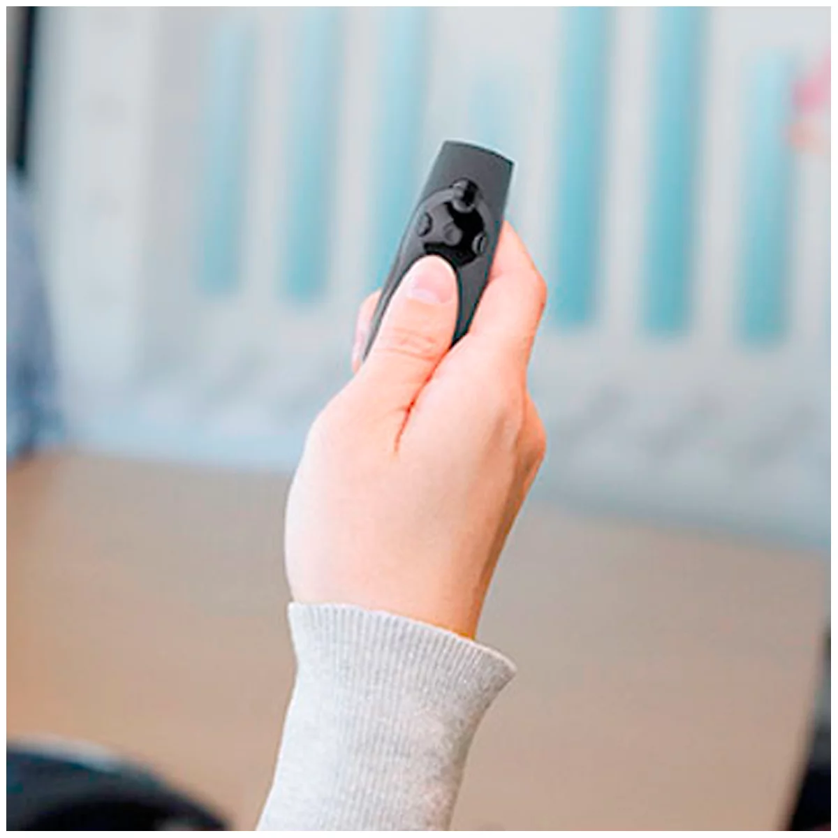 Personne tenant une télécommande noire avec des boutons. Pull gris. Environnement lumineux.