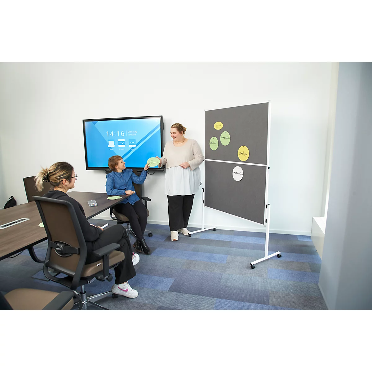 Three women in a conference room with presentation wall. One woman is presenting, two are listening.