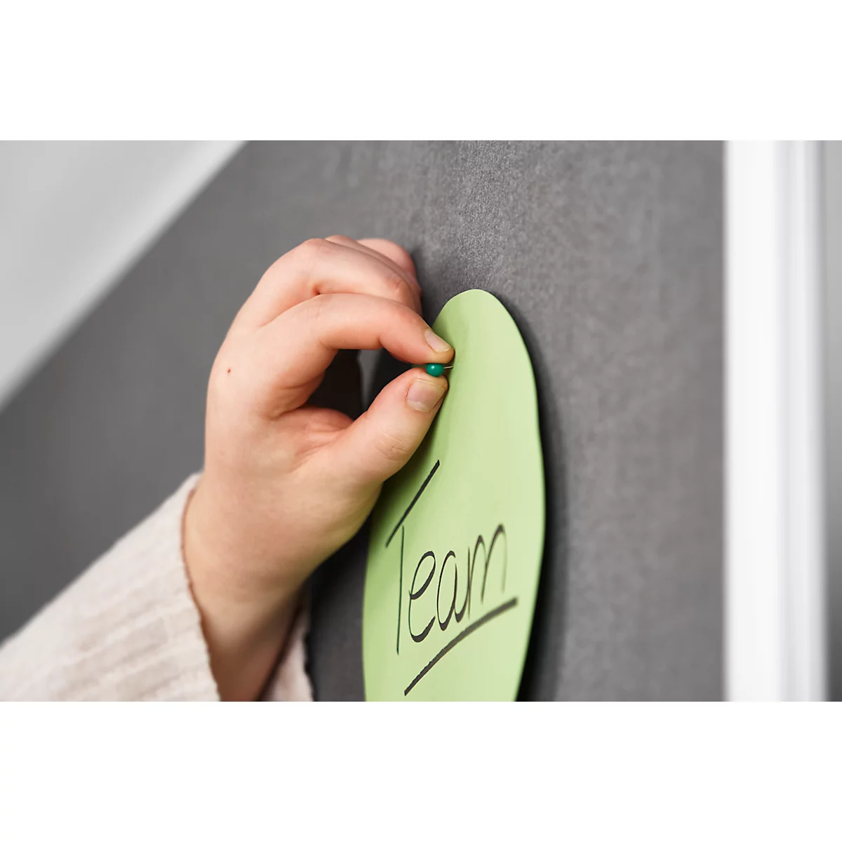 A hand presses a green pin onto a green leaf with the word Team on a dark bulletin board.