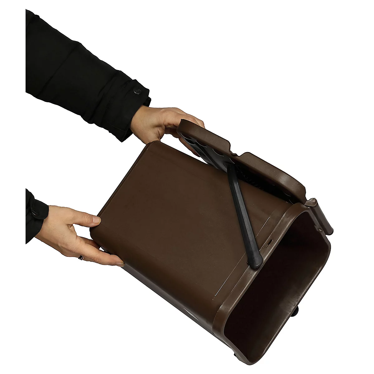 Persoon houdt bruine vuilnisbak vast, deksel open. Zwarte kleding, witte achtergrond.