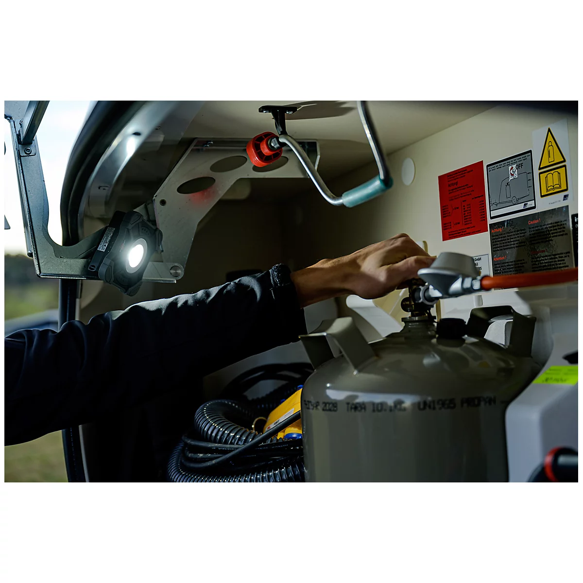 Une main ouvre la valve d'un cylindre de gaz gris dans un appareil technique. En arrière-plan, des avertissements et des tuyaux sont visibles.