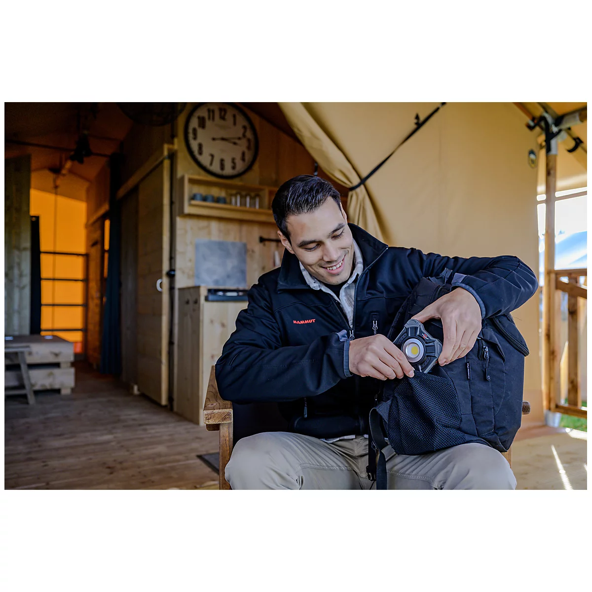 Un homme souriant porte une veste noire et est assis sur une chaise en bois. Il examine un sac à dos noir tout en fixant un objet rond et jaune dessus. En arrière-plan, on voit une cabane en bois avec une grande horloge et quelques appareils élect...