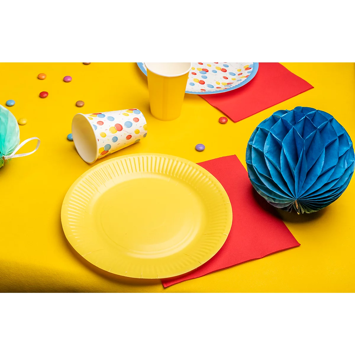 Assiette et gobelet jaunes, boule en papier bleue, serviettes rouges sur table jaune, bonbons, décoration de fête.