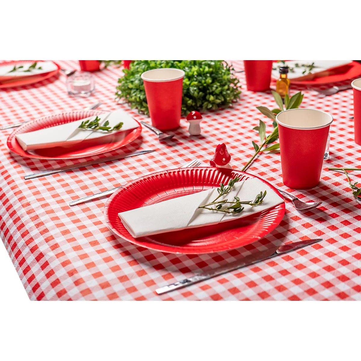 Une table dressée de façon festive avec une nappe à carreaux rouges et blancs. Assiettes, gobelets et serviettes rouges avec des brins d'herbes.