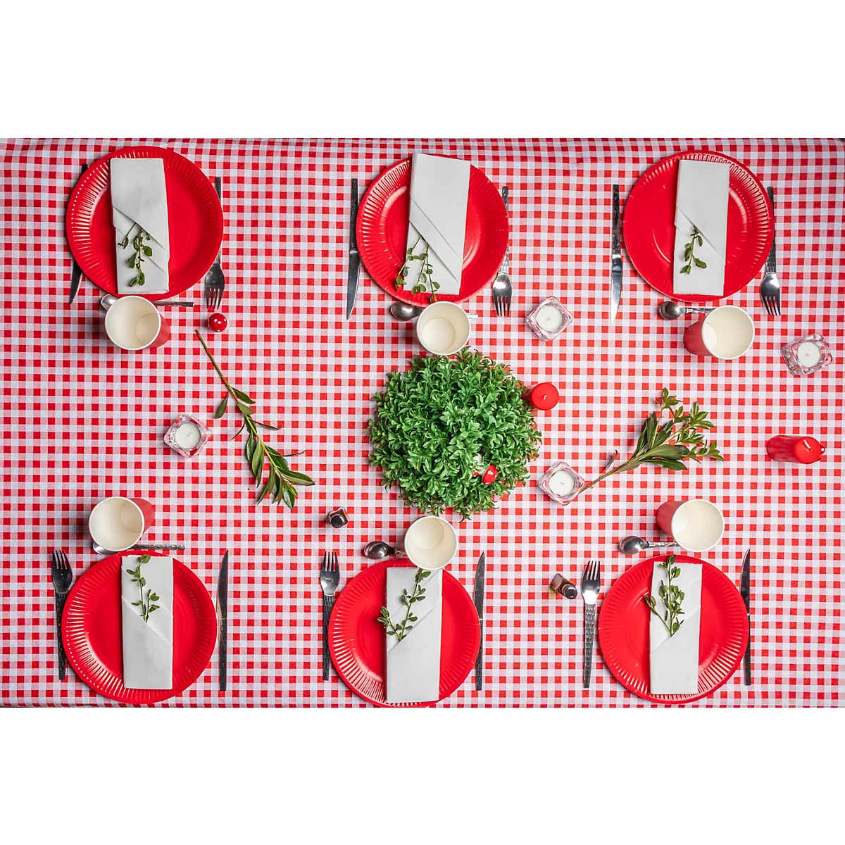 Table avec nappe à carreaux rouges. Assiettes rouges avec serviettes, couverts et tasses. Décoré de verdure et de bougies.