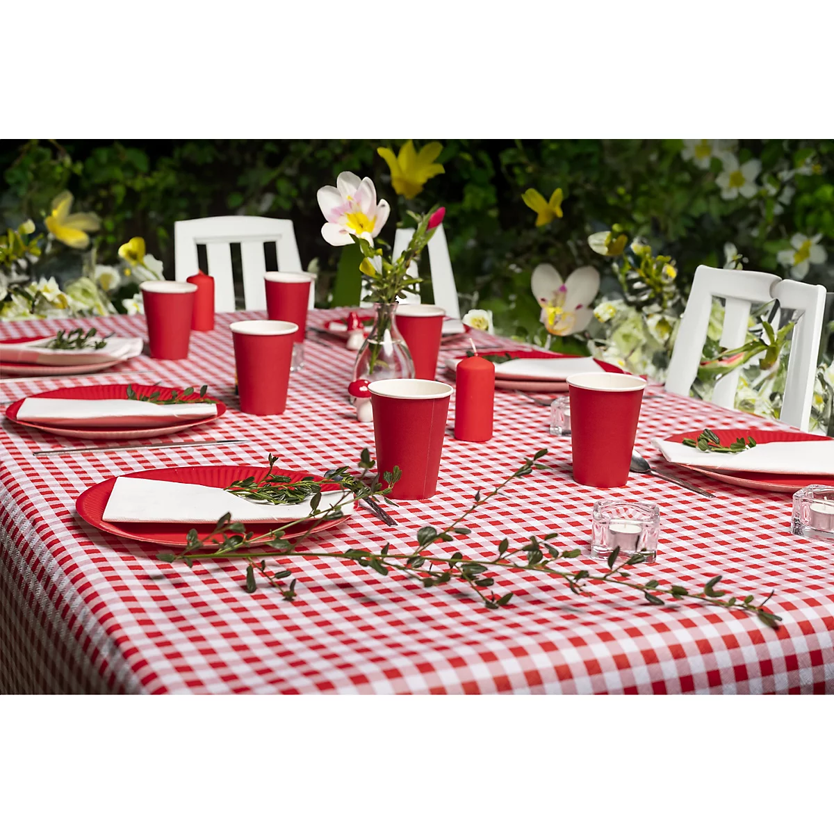 Ein festlich gedeckter Tisch mit rot-weiss karierter Tischdecke, roten Tellern, Bechern, Blumen und Kerzen im Freien.