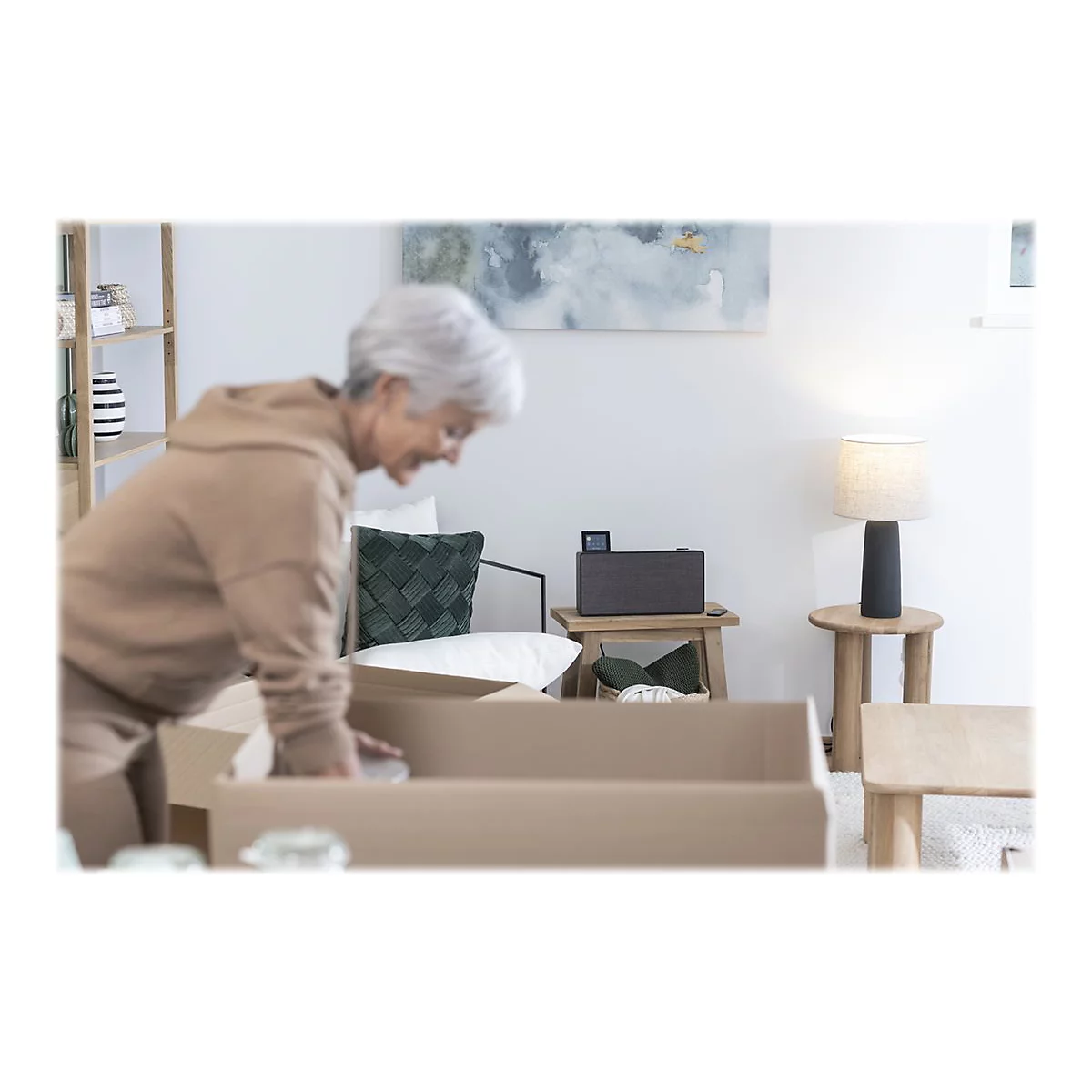 Ältere Frau packt Kisten aus. Im Hintergrund ein Lautsprecher, eine Lampe und ein Regal.