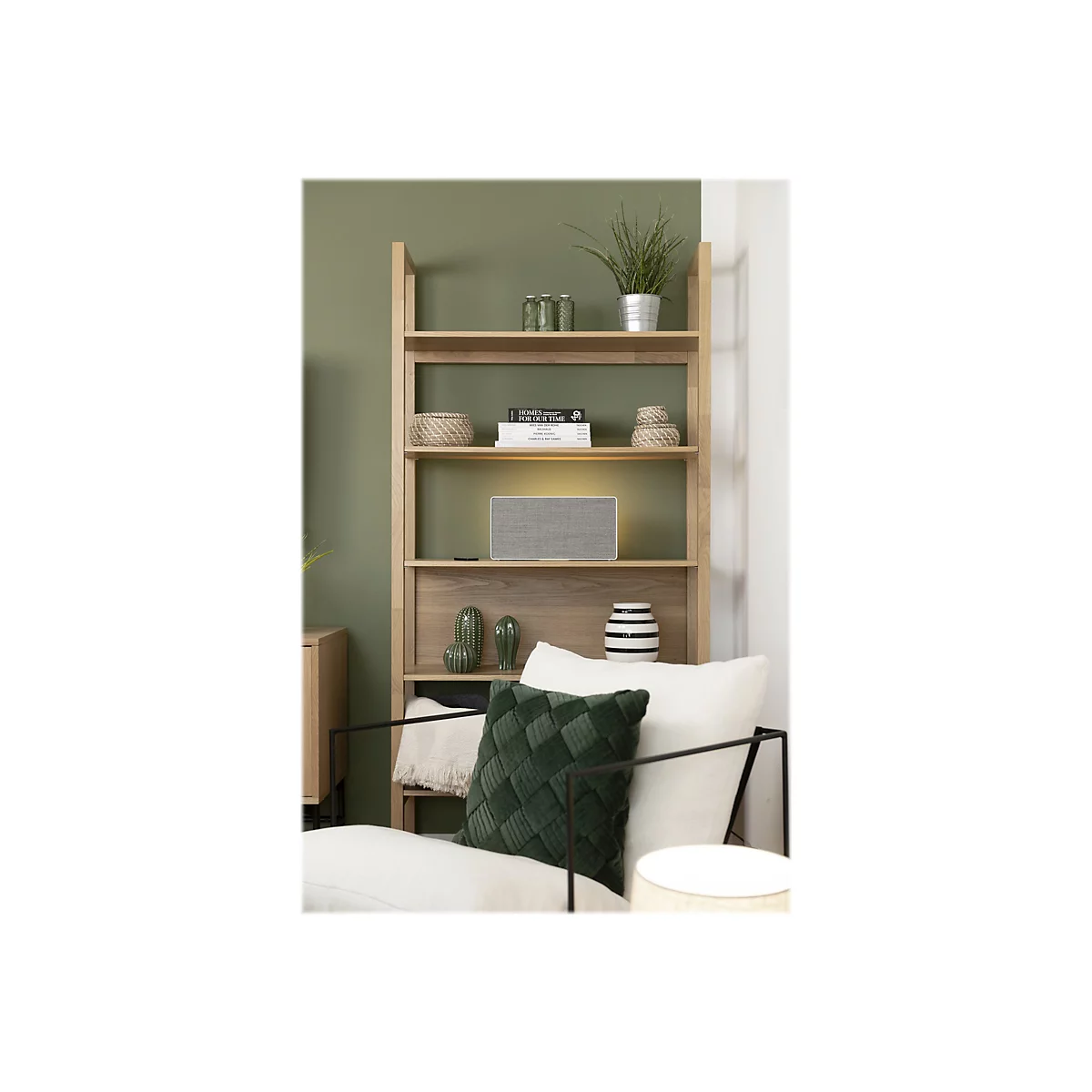 Ein stilvolles Wohnzimmer. Ein Holzregal, das mit Büchern, Dekorationen und einem grauen Lautsprecher bestückt ist, steht vor einer Wand in Salbeigrün. Ein grüner Samtsessel lädt zum Entspannen ein.