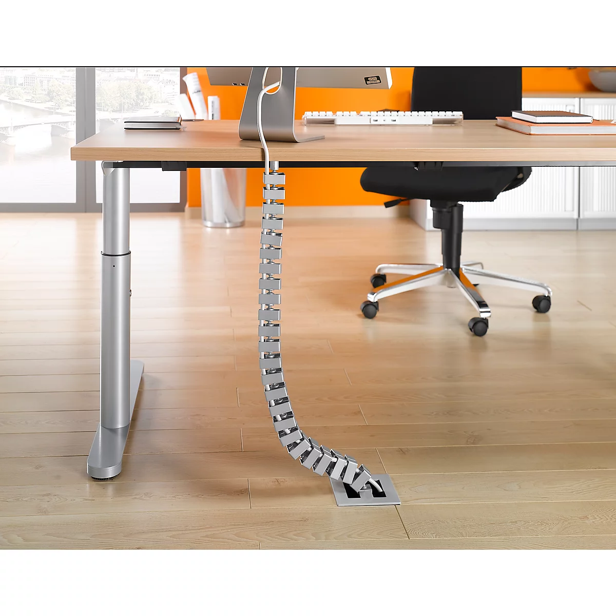 A metal cable duct on a height-adjustable desk. There is a monitor, keyboard and books on the table.