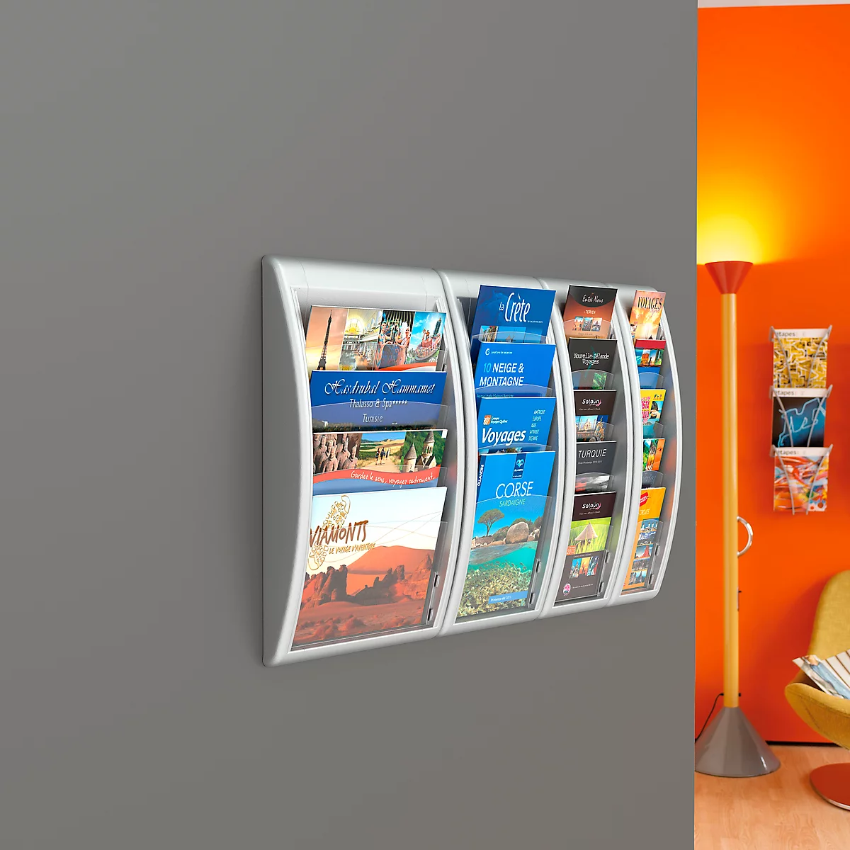 White curved magazine rack with brochures on grey wall. In the background, an orange wall section with floor lamp.