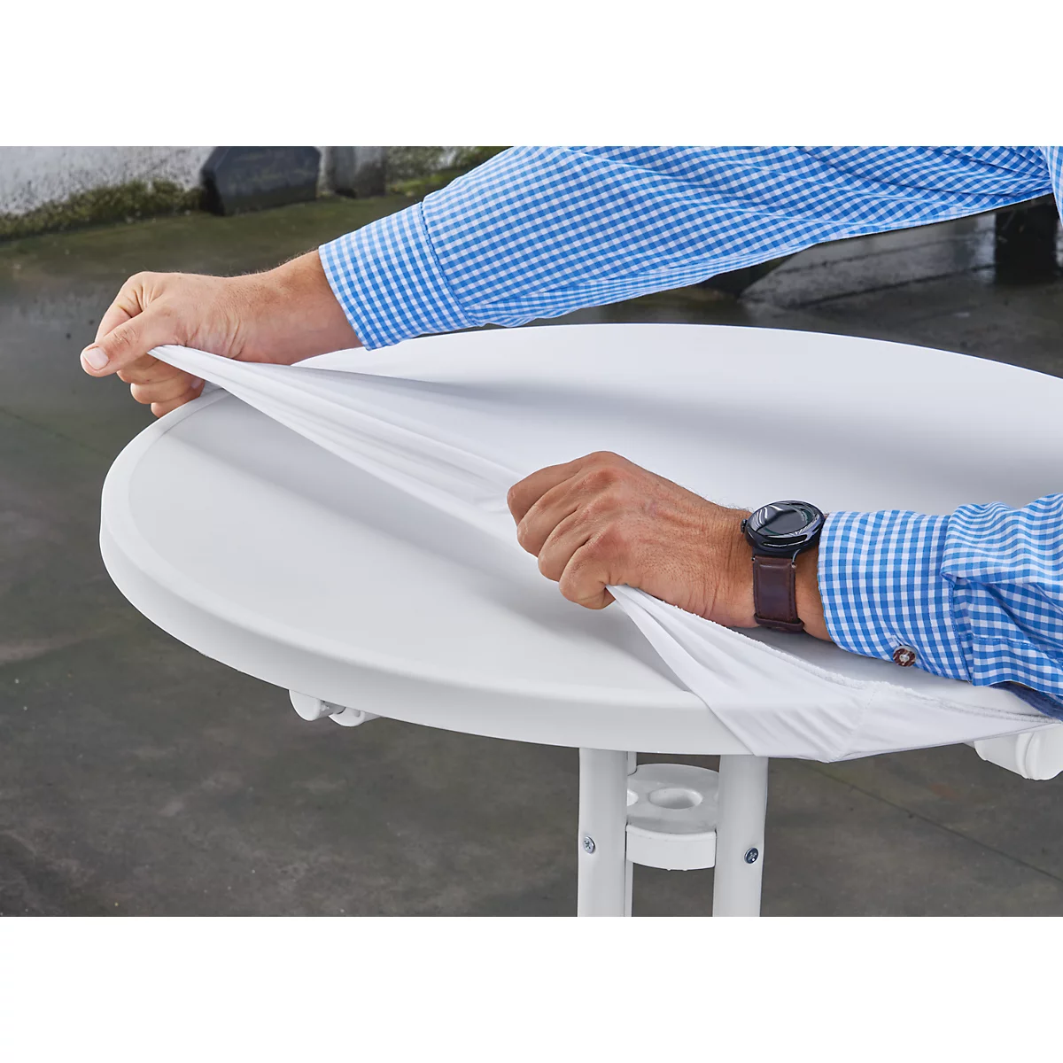 Hands pulling white fabric off round table. Wristwatch visible. Blurred background.