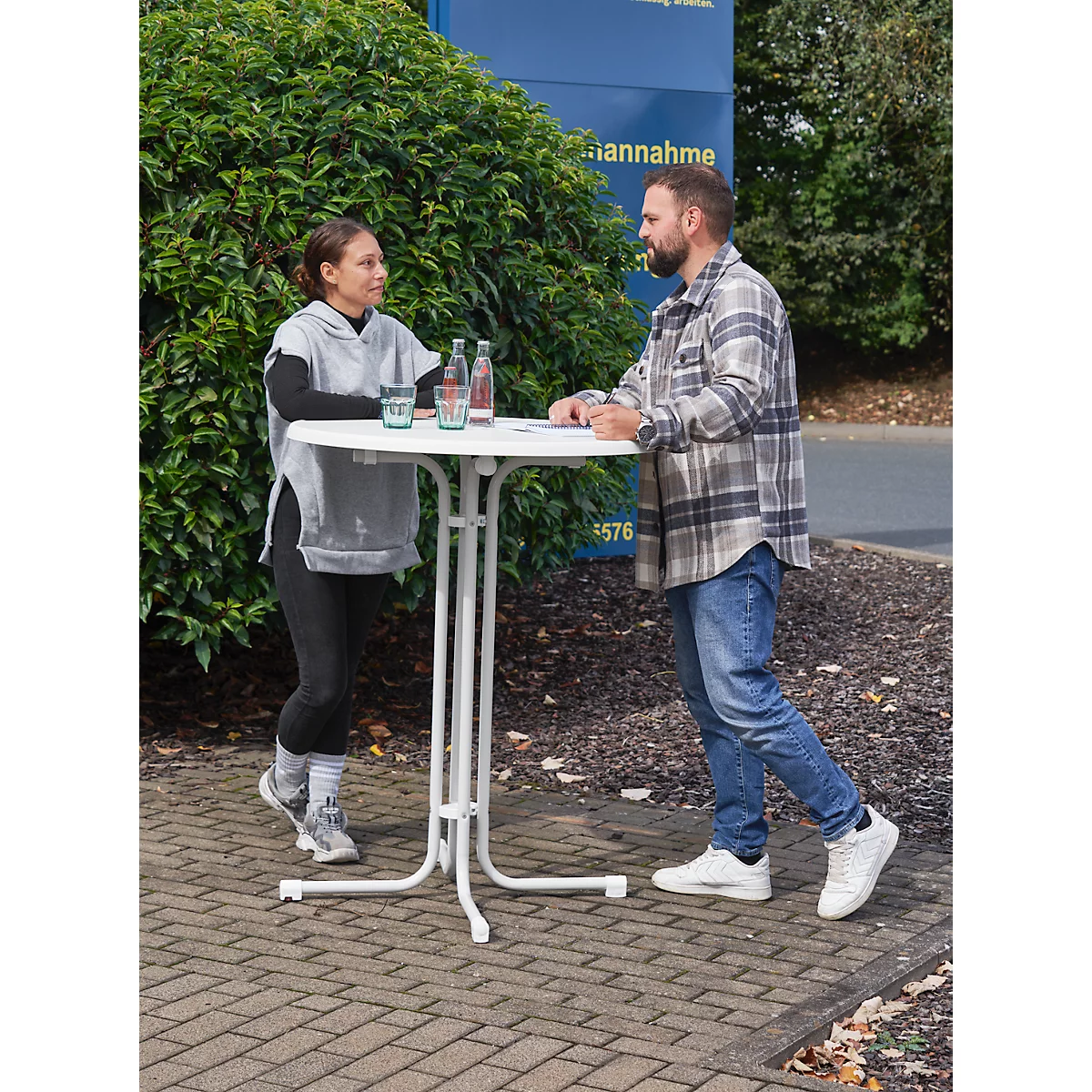 Two people standing at a white high table outdoors. Drinks are on the table. A woman and a man are talking.