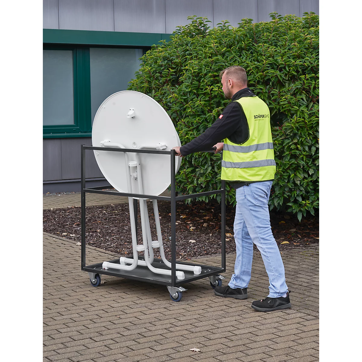 A man in a safety vest pushes a serving cart with a round, white table.
