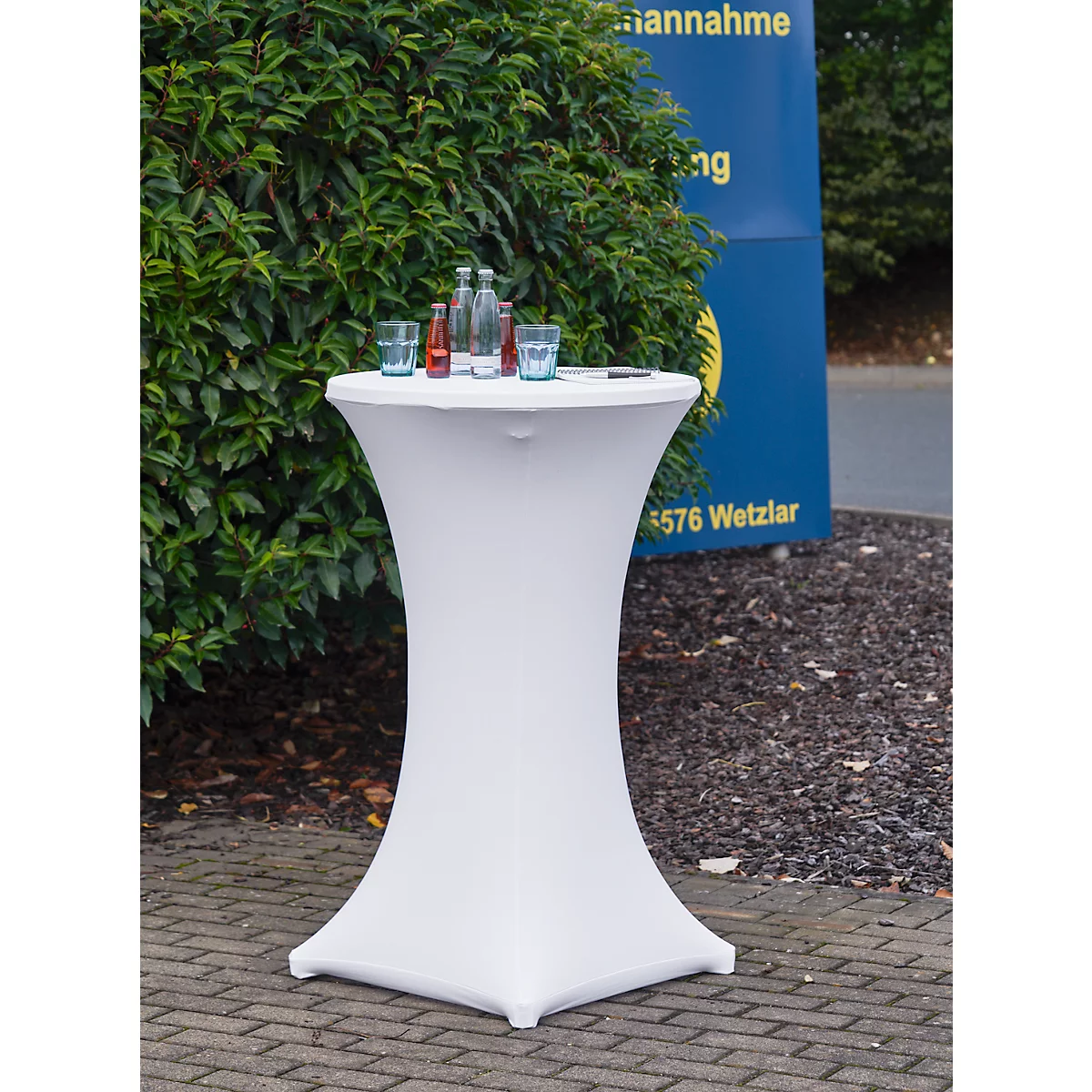 Witte statafel met drankjes, voor een heg en een blauw bord.