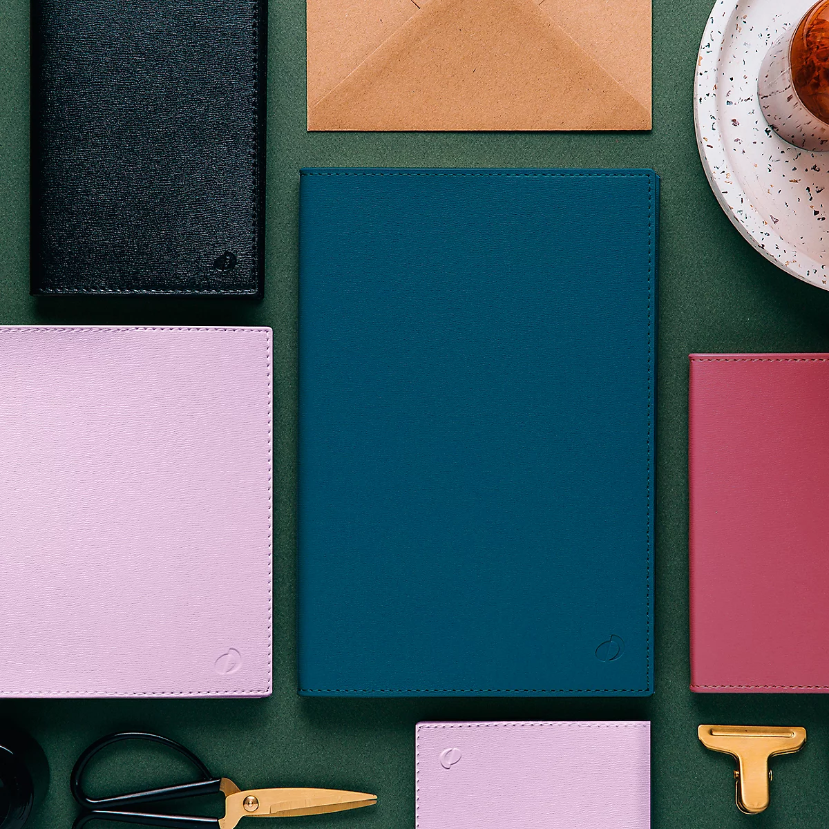 Draufsicht von verschiedenen Lederwaren in verschiedenen Farben: ein blaues Notizbuch, eine rosa Hülle, eine dunkle Brieftasche. Auf grünem Hintergrund.