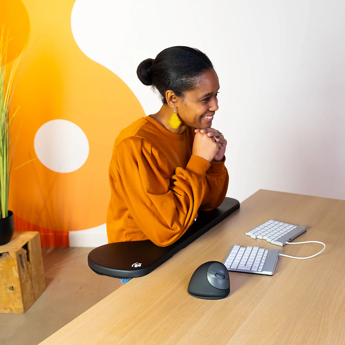 A smiling woman sits at a desk. She wears an ochre sweater and yellow earrings. Keyboards and an ergonomic mouse are on the desk.