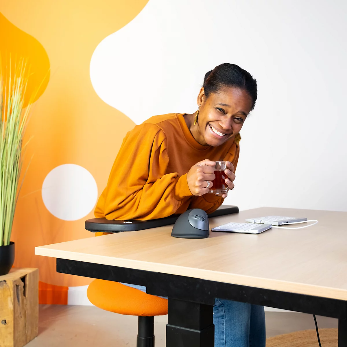 A smiling woman drinks tea at a desk. She is wearing an orange top and sitting at a standing desk.