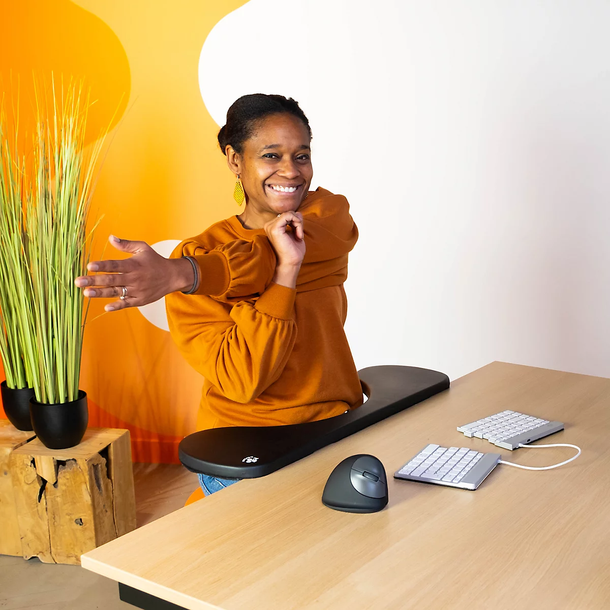 Smiling woman stretching at a desk, ergonomic accessories in front: mouse, keyboard, tray.