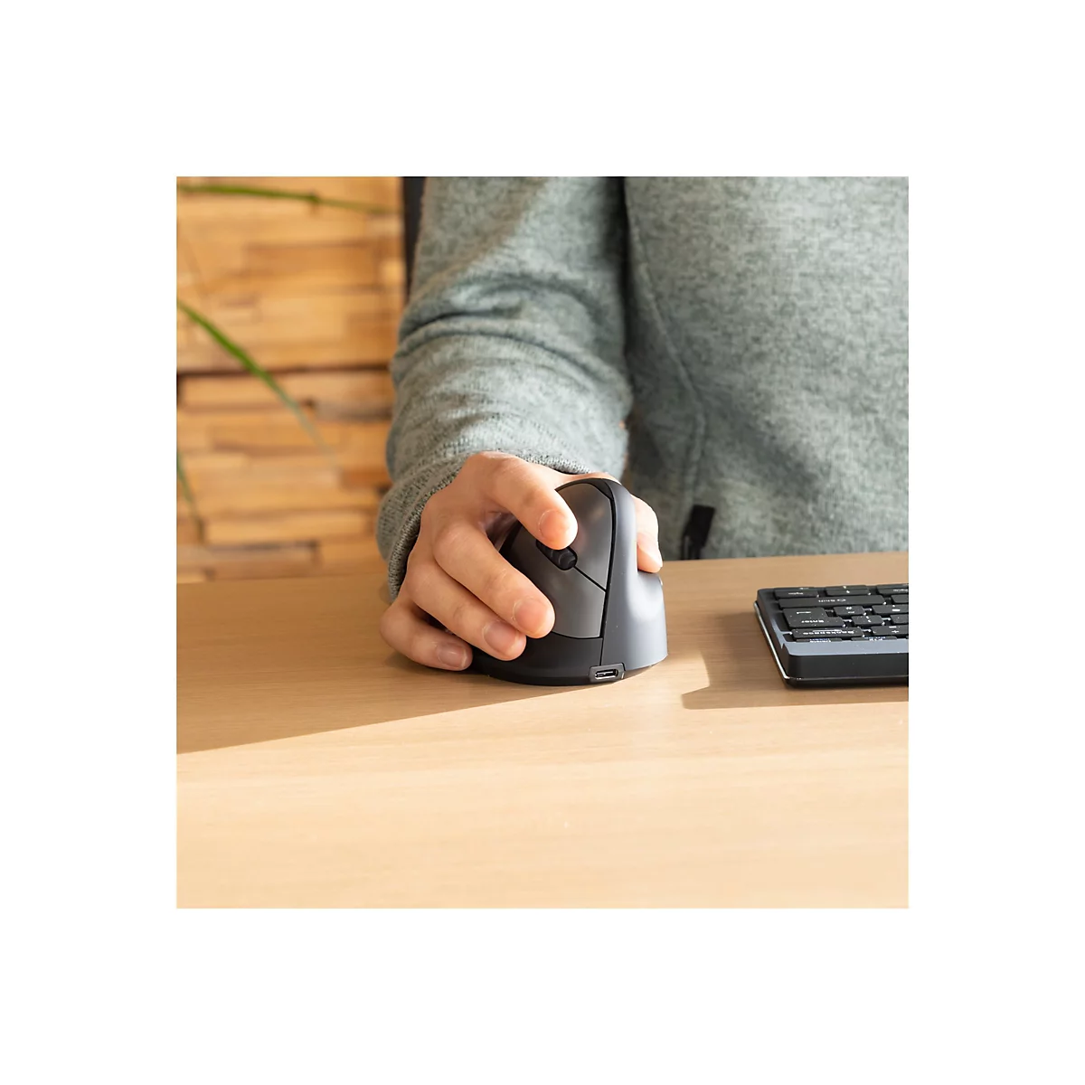 Person benutzt eine ergonomische Maus. Graue Maus auf Holzschreibtisch, neben Tastatur. Hintergrund: Holzwand.