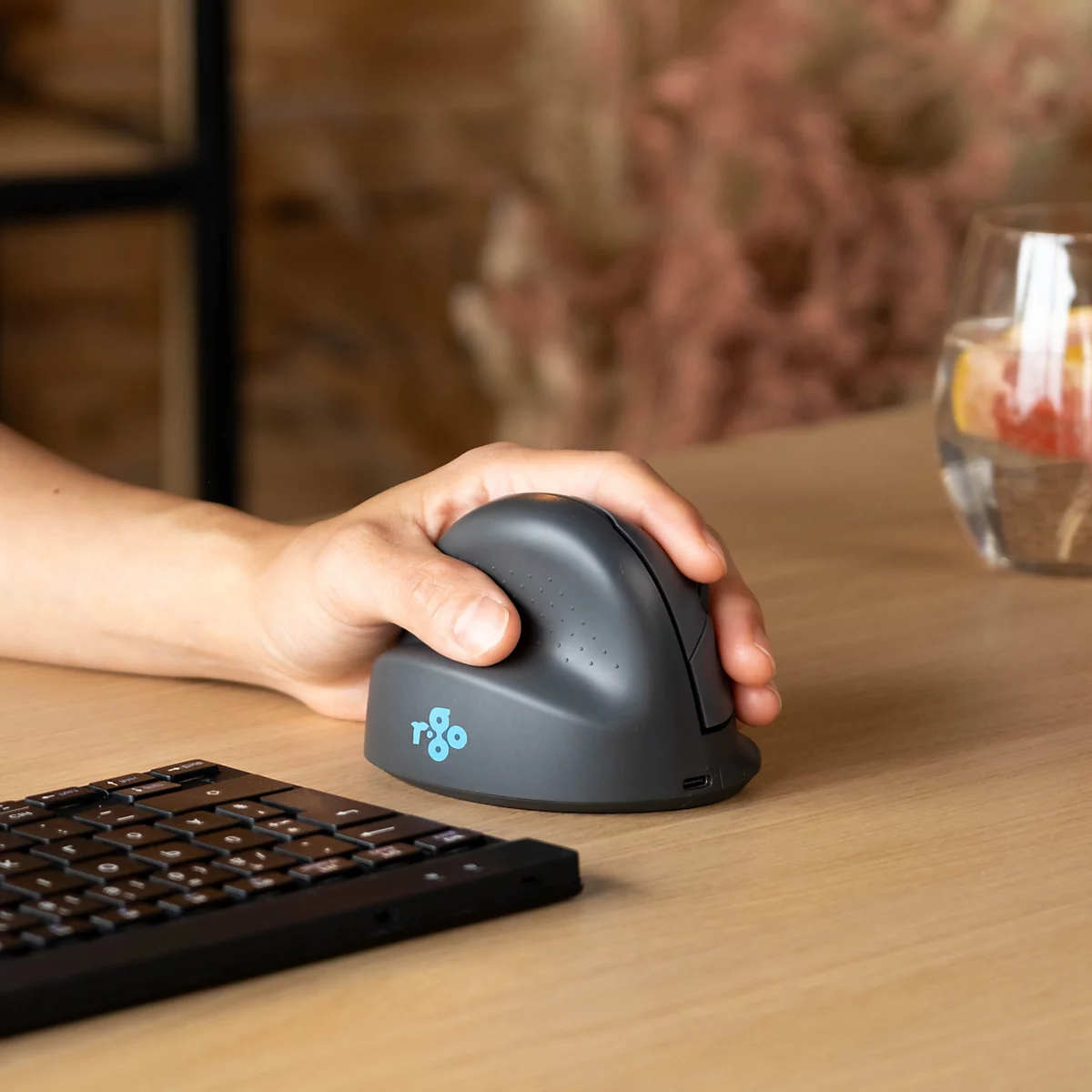Hand bedient vertikale, anthrazitfarbene ergonomische Maus auf einem Holztisch neben einer Tastatur.
