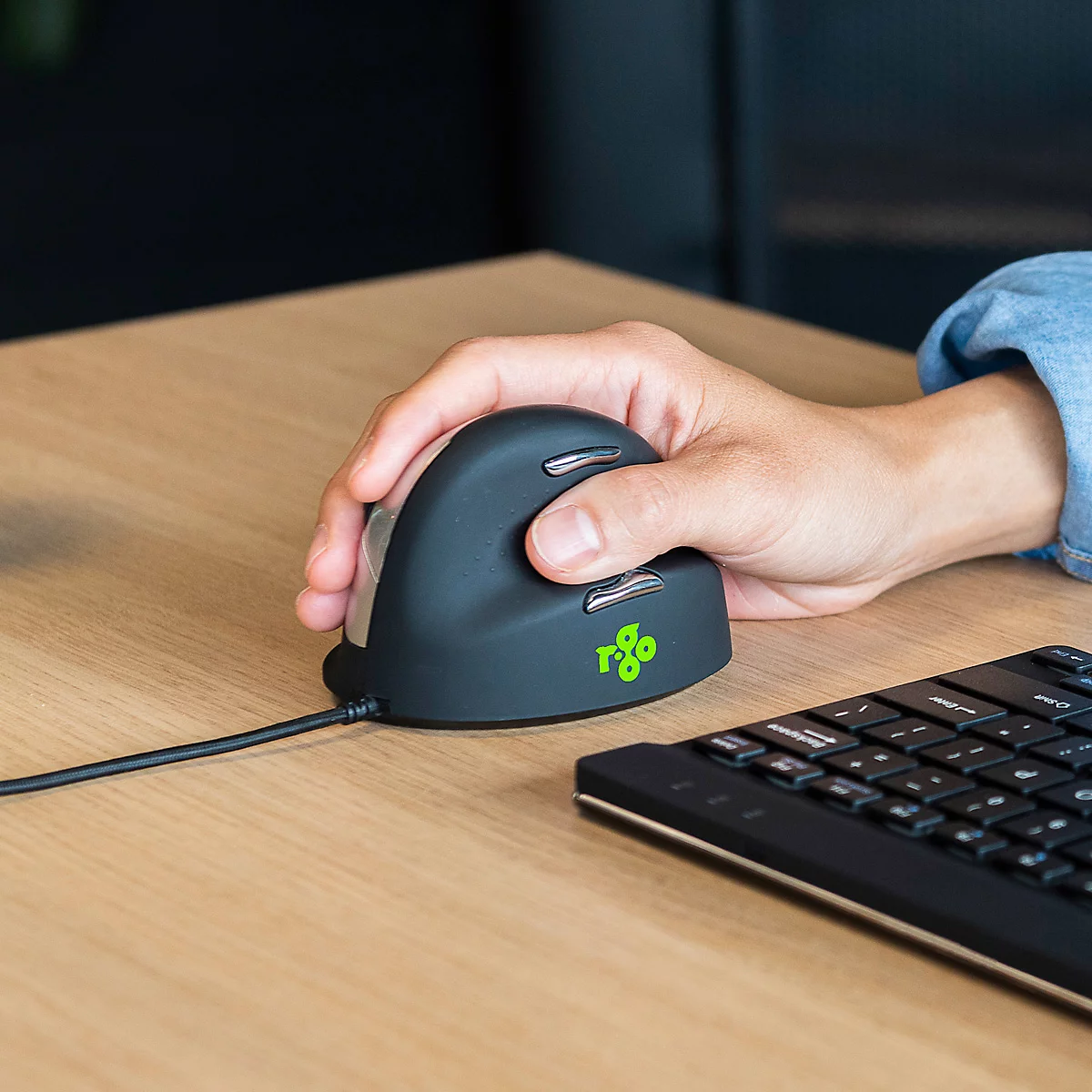 Hand greift ergonomische Maus. Auf dem Tisch liegen zudem eine Tastatur und ein Kabel.