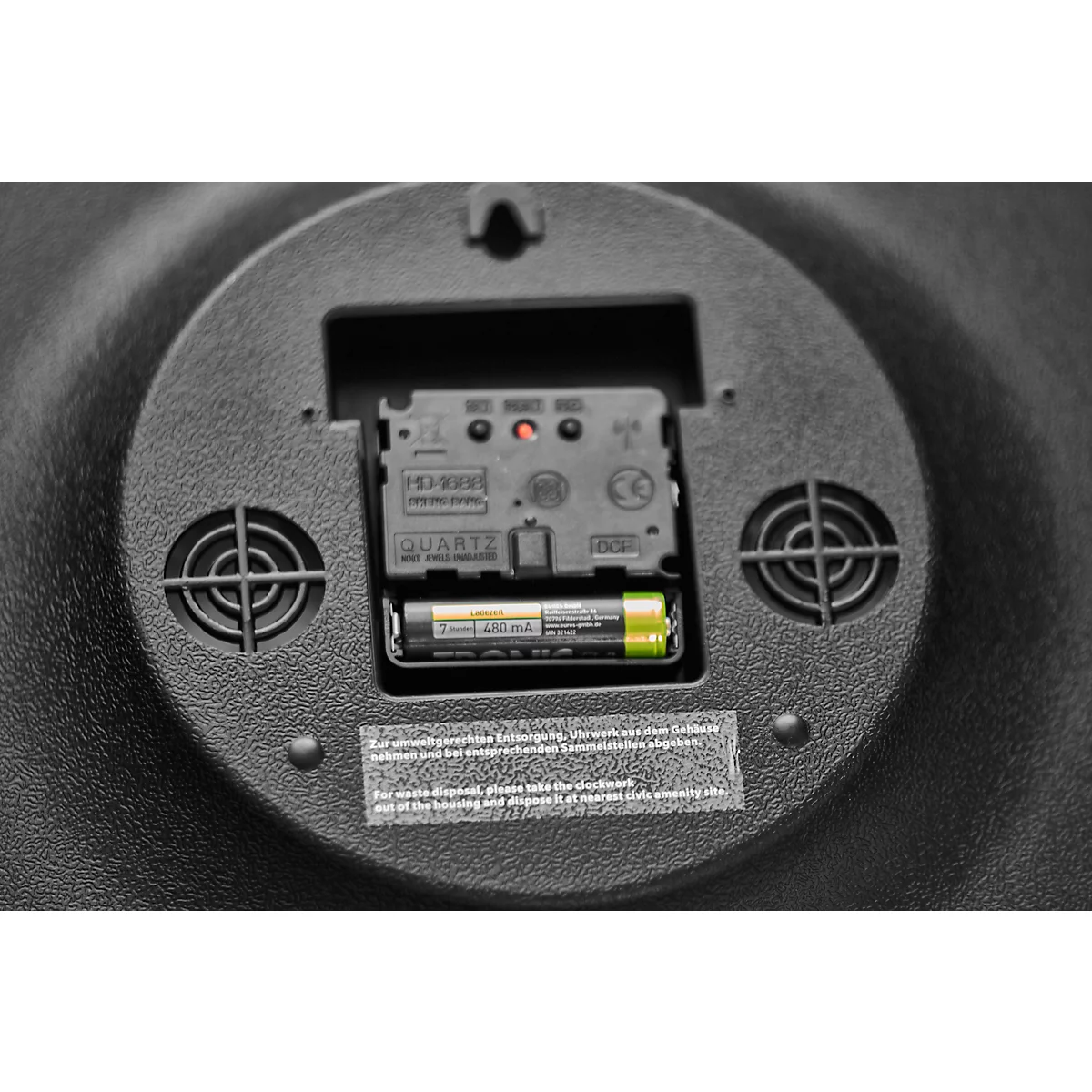 Close-up of the back of a clock with a battery installed. Black casing, ventilation slots, battery and clock mechanism visible.