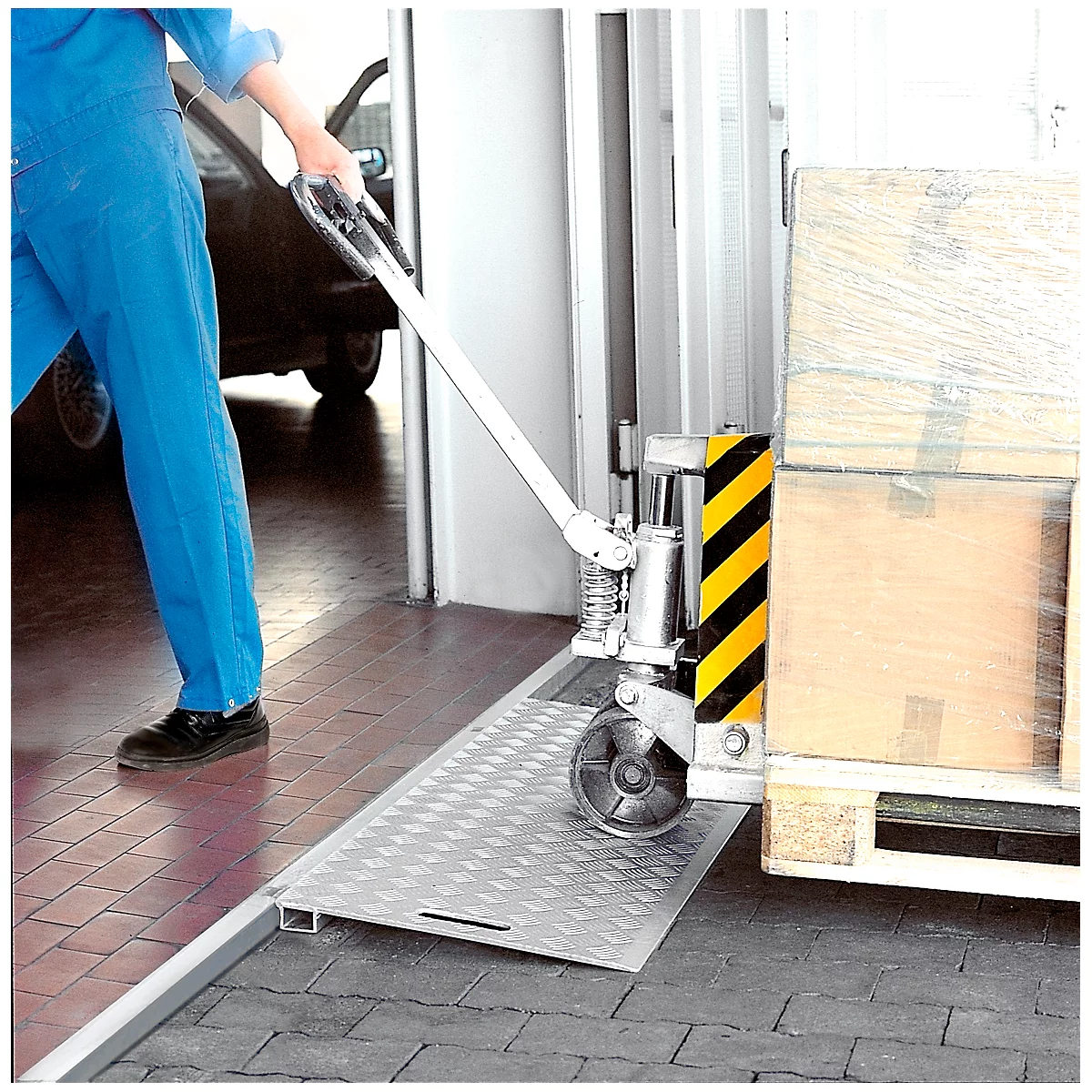 Un homme en bleu de travail utilise une rampe de chargement avec un transpalette. Des palettes sont visibles en arrière-plan.