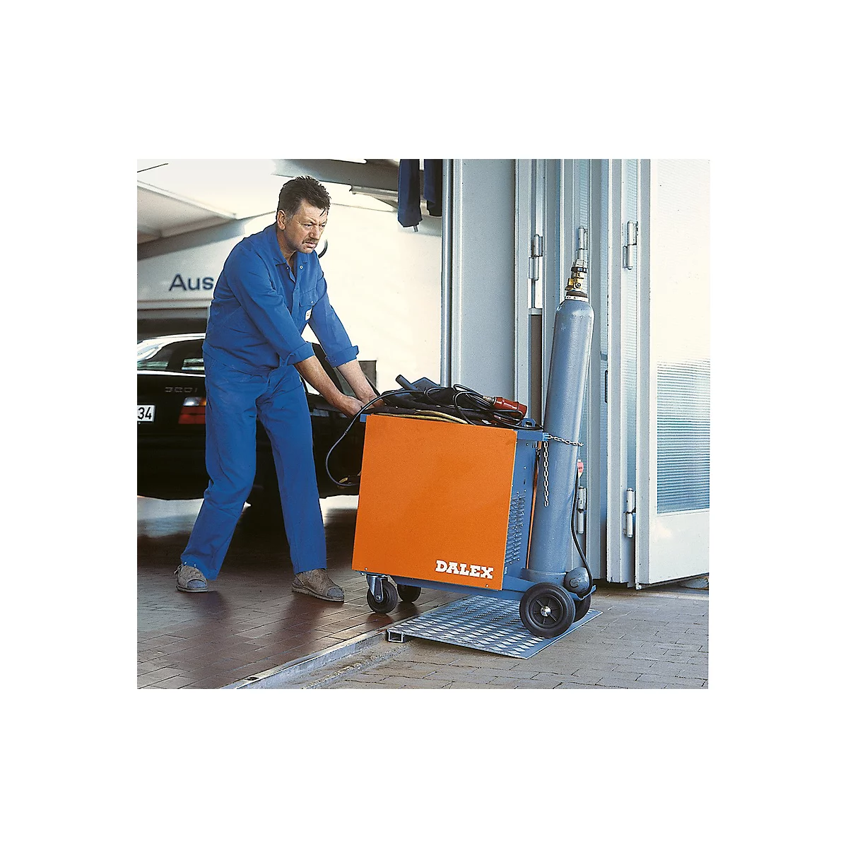 Un homme en salopette bleue pousse un chariot de soudage avec un coffre orange et une bouteille de gaz.