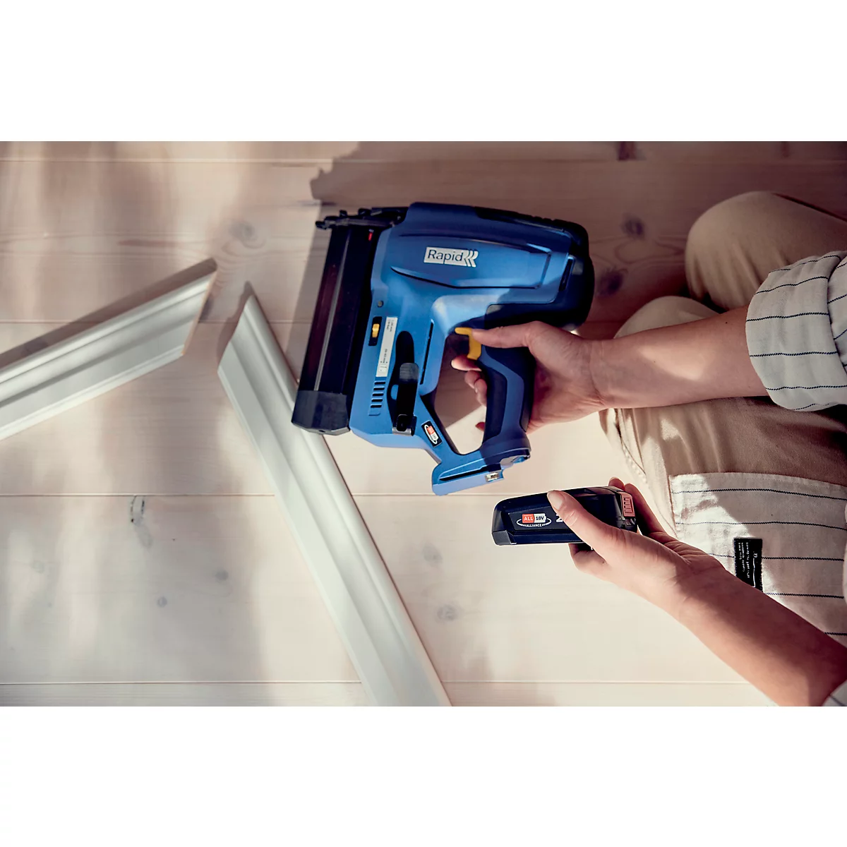 Een persoon houdt een blauwe nietmachine en een batterij vast. Lijsten en een houten oppervlak op de achtergrond.