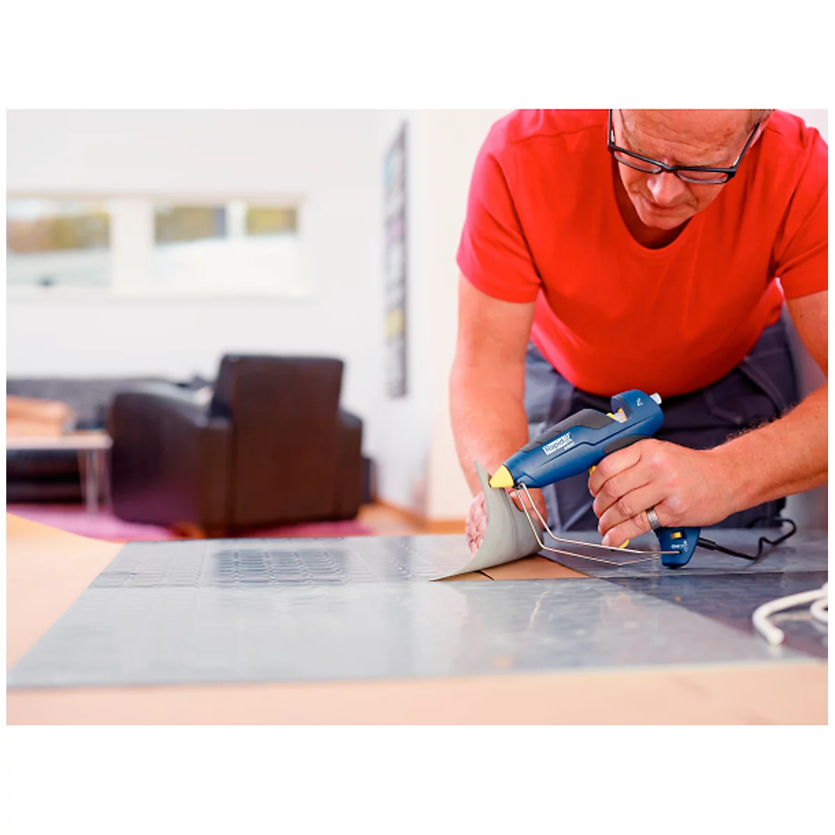 A man glues something onto a plate with a hot glue gun. He is wearing a red t-shirt and glasses.