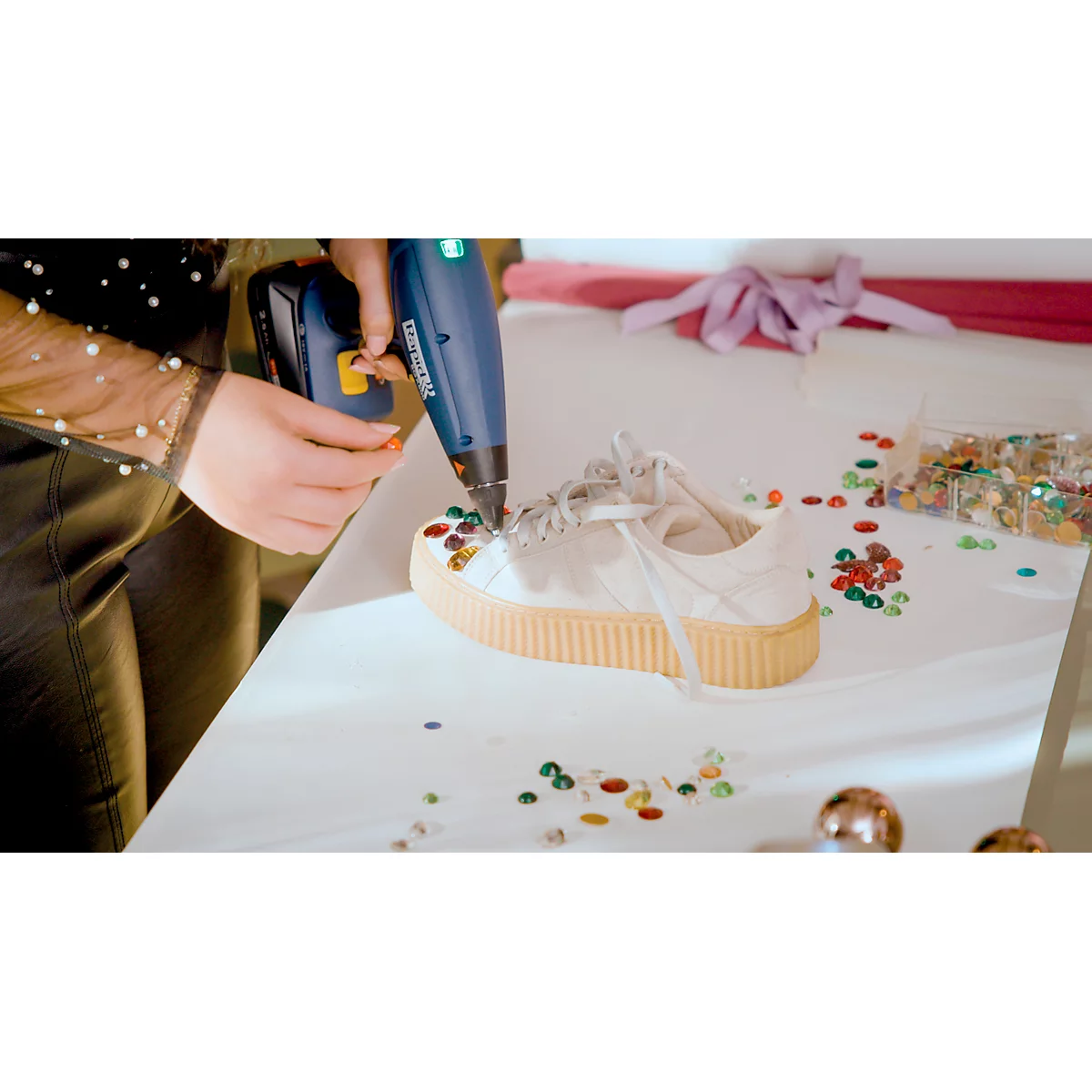 A person is gluing rhinestones onto a white shoe with a hot glue gun.