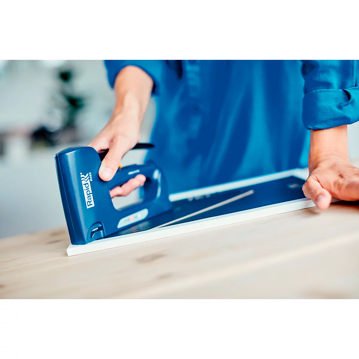 A person is attaching a white object to a wooden surface with a blue stapler. The stapler has the word "Rapid" on it.