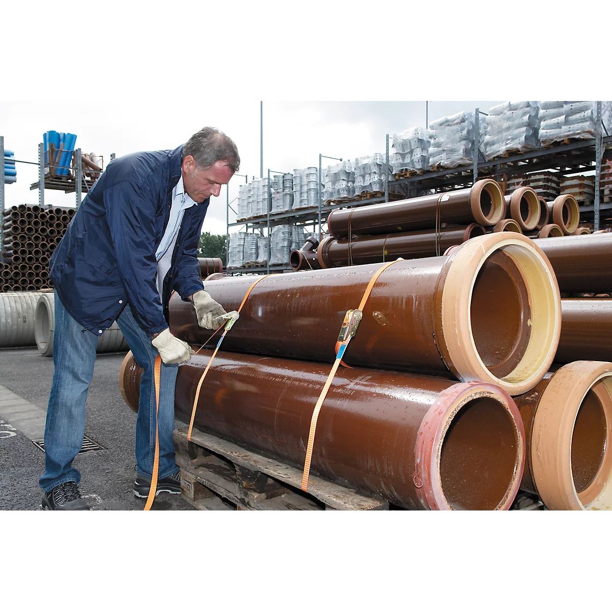 A man in a blue jacket secures brown pipes with straps. In the background, shelves with more pipes.