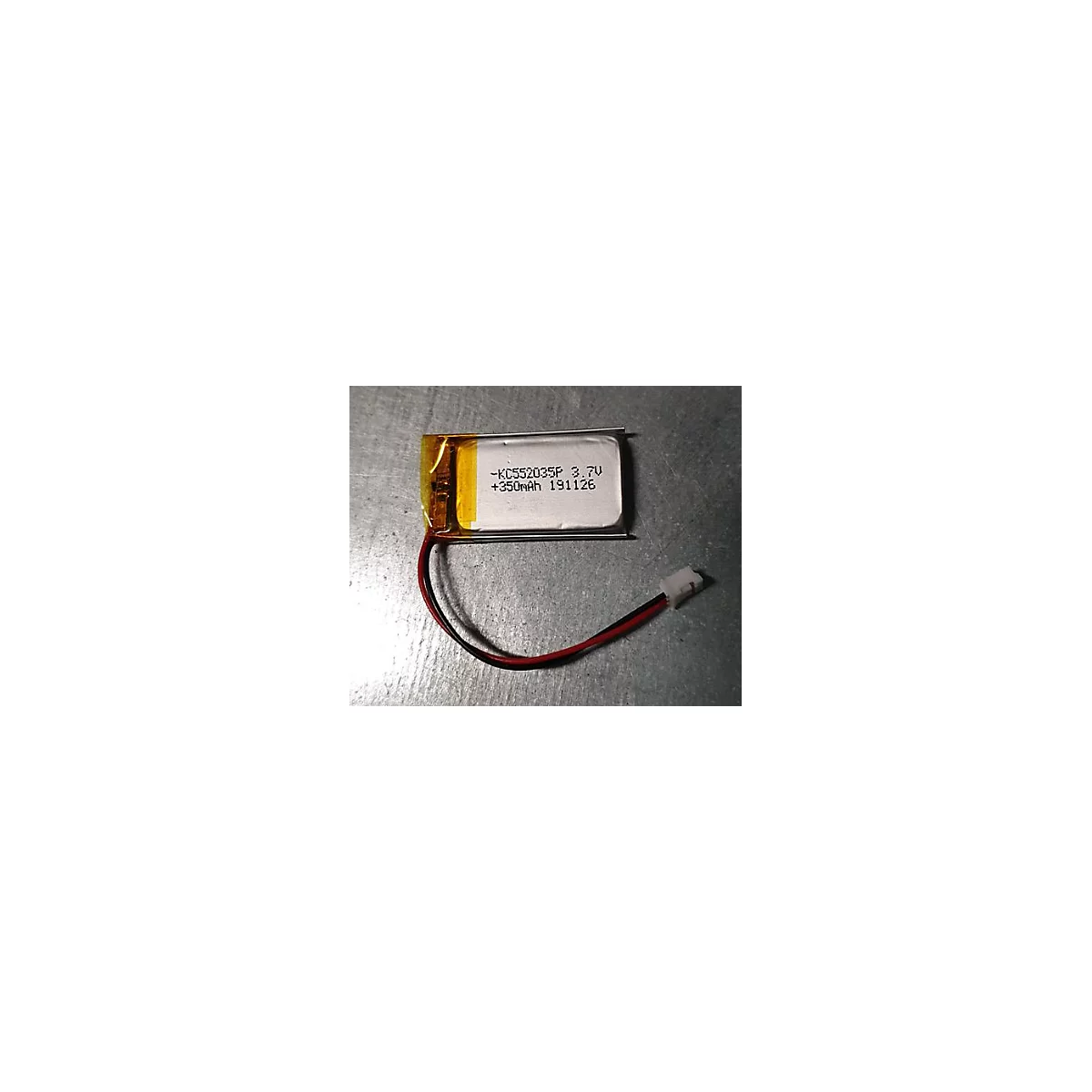 Rechteckiger Lithium-Polymer-Akku mit gelber Isolierung, Kabeln und weißem Stecker auf metallischem Untergrund.