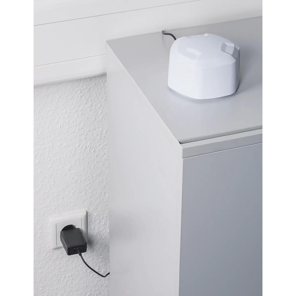 Ein weißer, kompakter Sensor steht auf einem grauen Schrank und ist per Kabel mit einer Steckdose verbunden.
