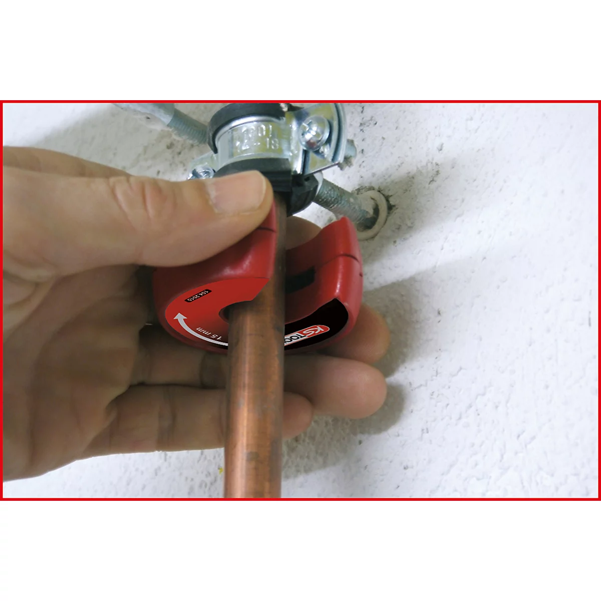 Hand hält ein rotes Werkzeug, das ein Kupferrohr schneidet. Auf dem Rohr steht „15mm“ und ein Pfeil.
