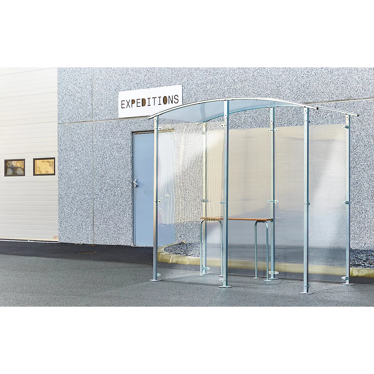 Heller, transparenter Unterstand mit Metallrahmen und Holztisch vor einer Lagerhalle mit Schild "Expeditionen".