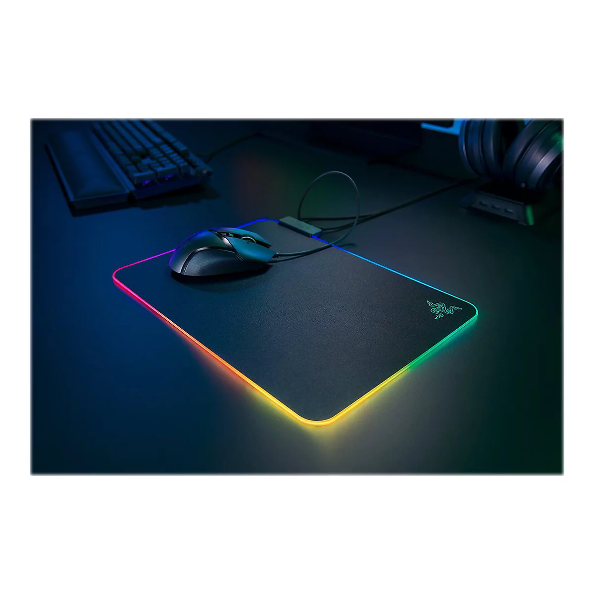 Schwarzes Gaming-Mauspad mit mehrfarbiger LED-Beleuchtung. Eine Maus liegt auf dem Pad, eine Tastatur und ein Headset sind im Hintergrund zu sehen.