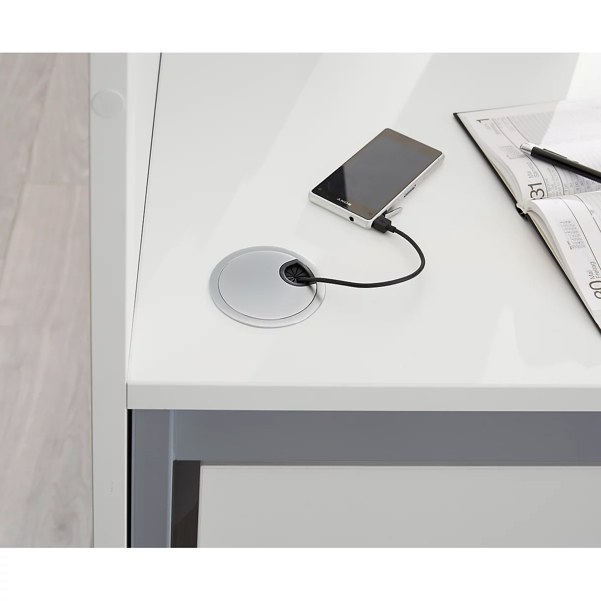 White desk with cable management. A smartphone is charging. A notebook and pen are lying next to it.