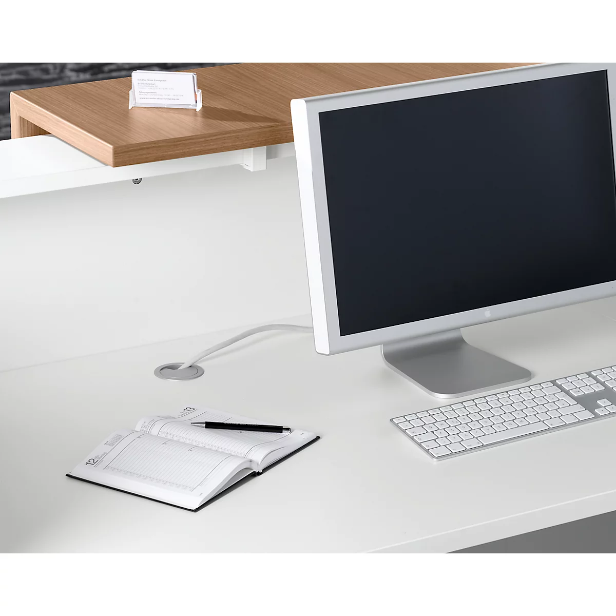 Desk with monitor, keyboard, notebook, and pen. A business card rests on a shelf.