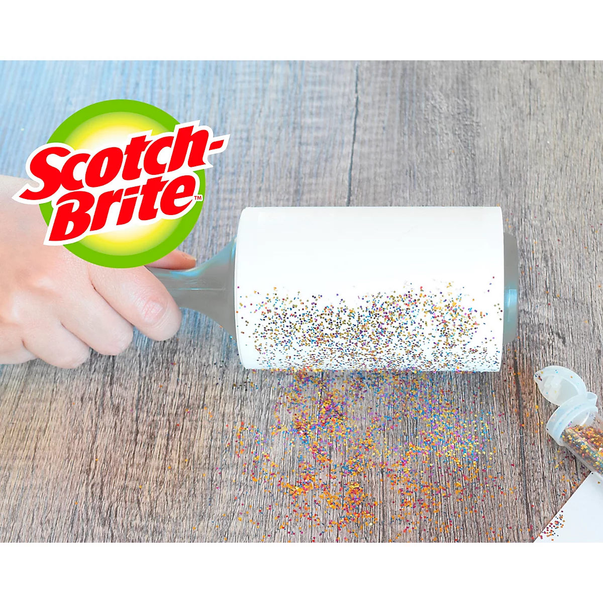 Un rouleau anti-peluches retire des paillettes d'une surface en bois. À côté se trouve un récipient contenant des paillettes. Le logo Scotch-Brite se trouve en haut à gauche.