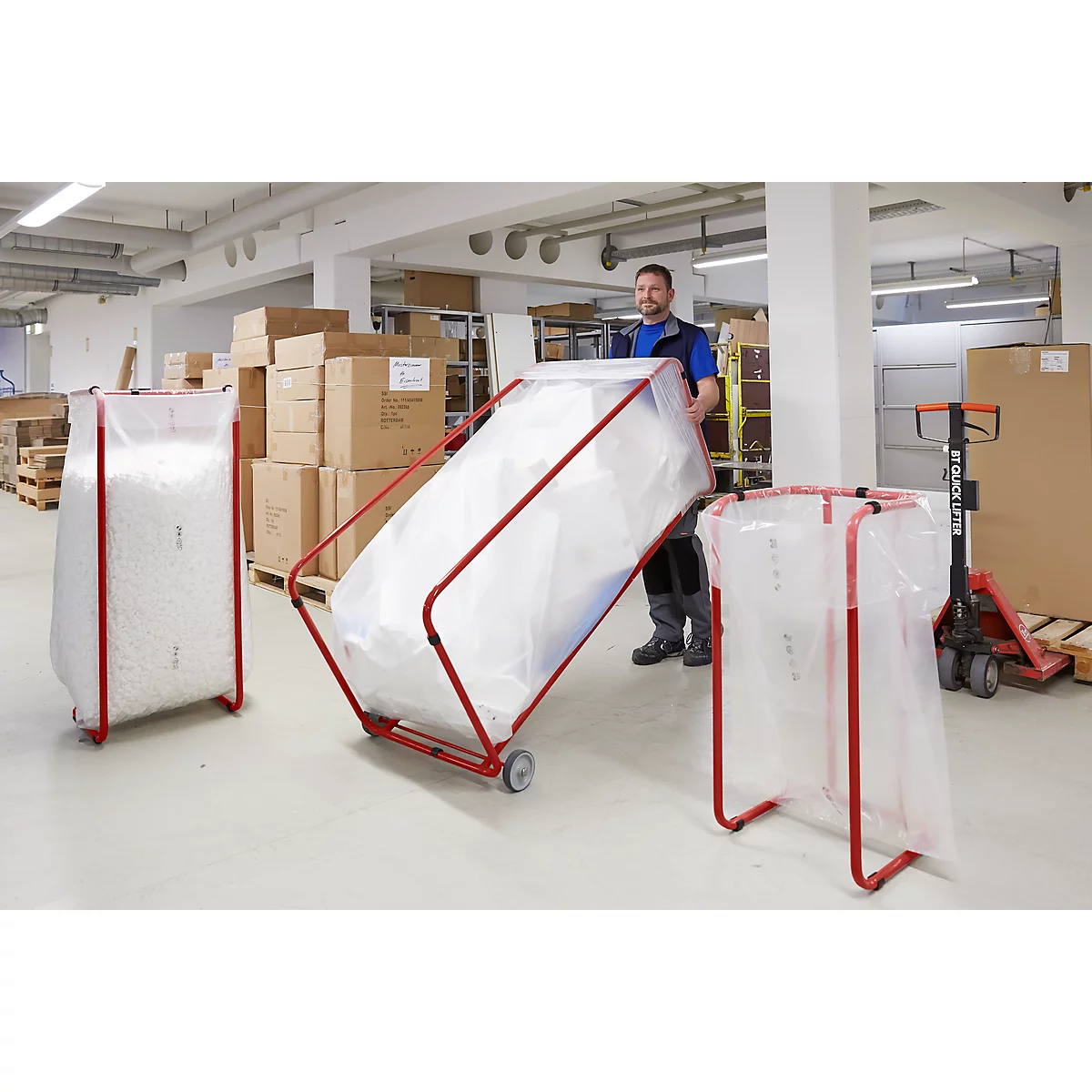 A man pushing a red trolley with a large white bag through a warehouse. Two more bags are standing next to it.