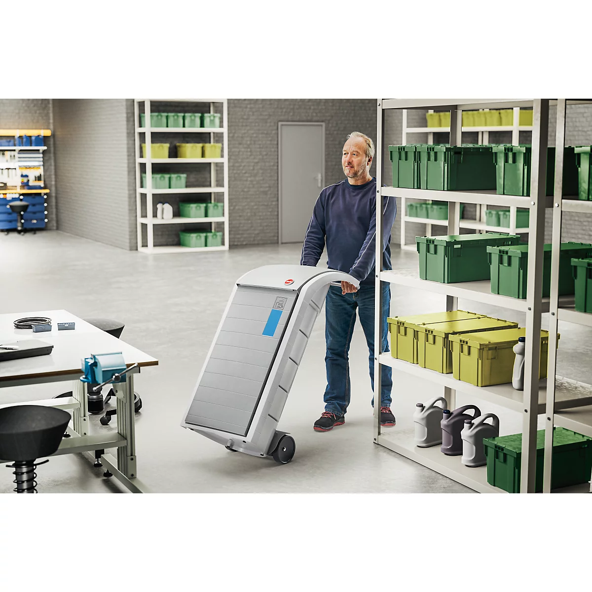 A man pulls a gray transport cart through a warehouse. Shelves with boxes are in the background.