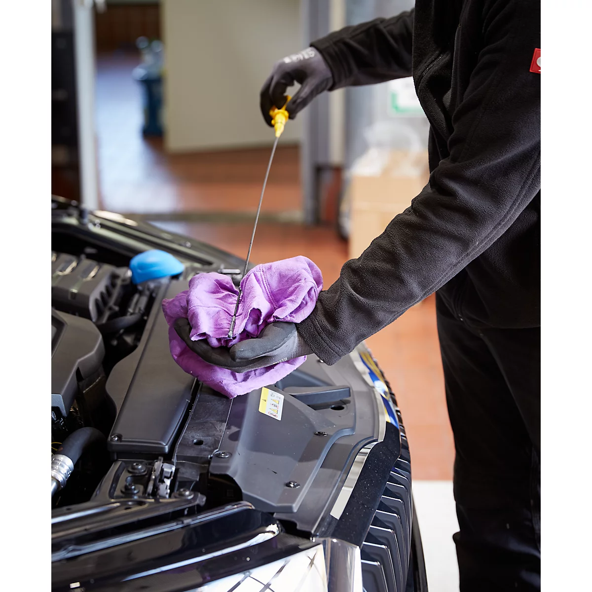 Een monteur controleert het oliepeil van een auto. Hij houdt een oliepeilstok en een paarse doek vast.
