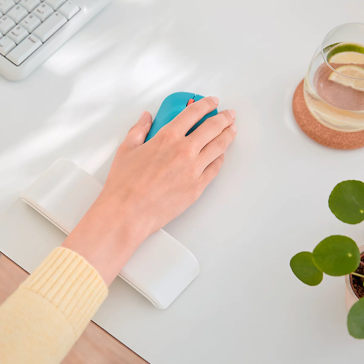 Main utilisant une souris bleue sur un bureau blanc, avec clavier, verre de boisson et plantes.