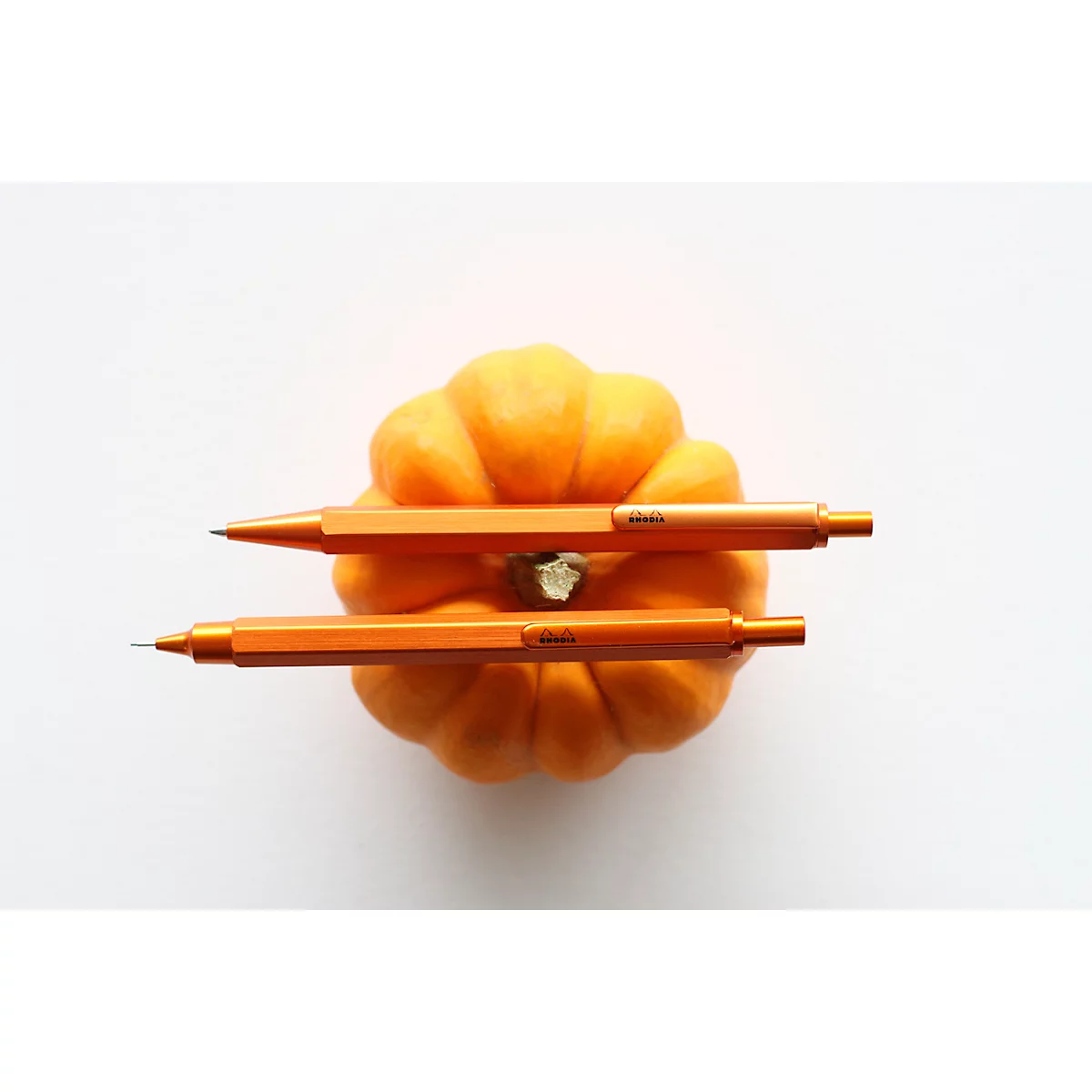 Zwei orangefarbene Bleistifte liegen auf einem kleinen Kürbis auf weissem Hintergrund.