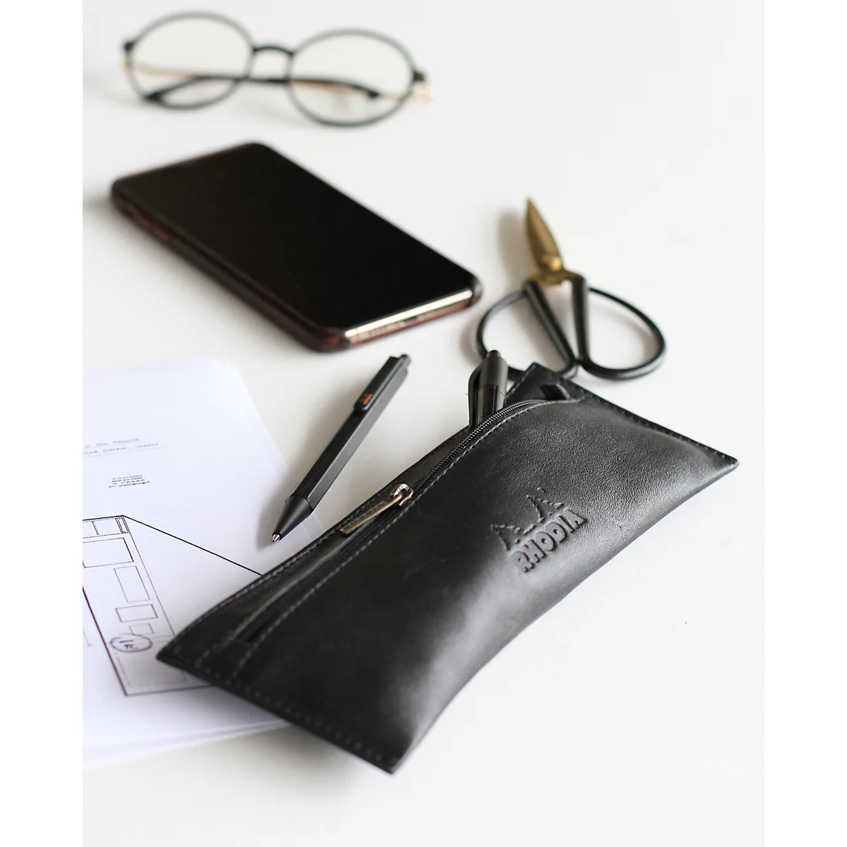 Schwarzes Etui mit Reissverschluss, daneben Brille, Smartphone, Schere, Stift und Papiere auf hellem Untergrund.
