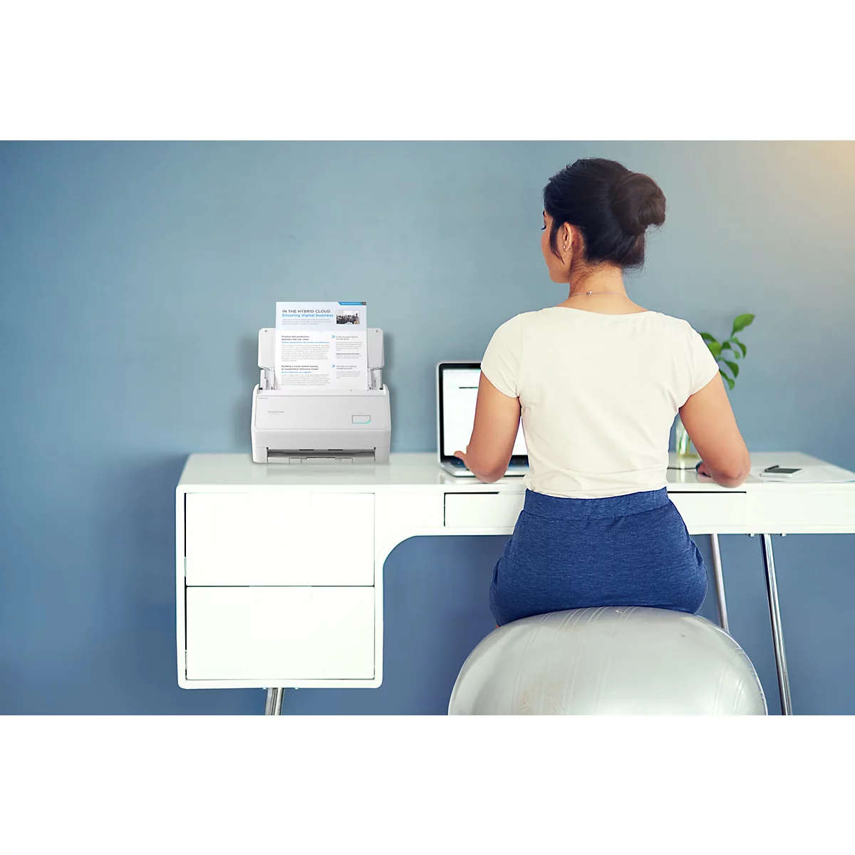 Eine Frau arbeitet an einem Schreibtisch mit Laptop und Dokumentenscanner vor einer blauen Wand.